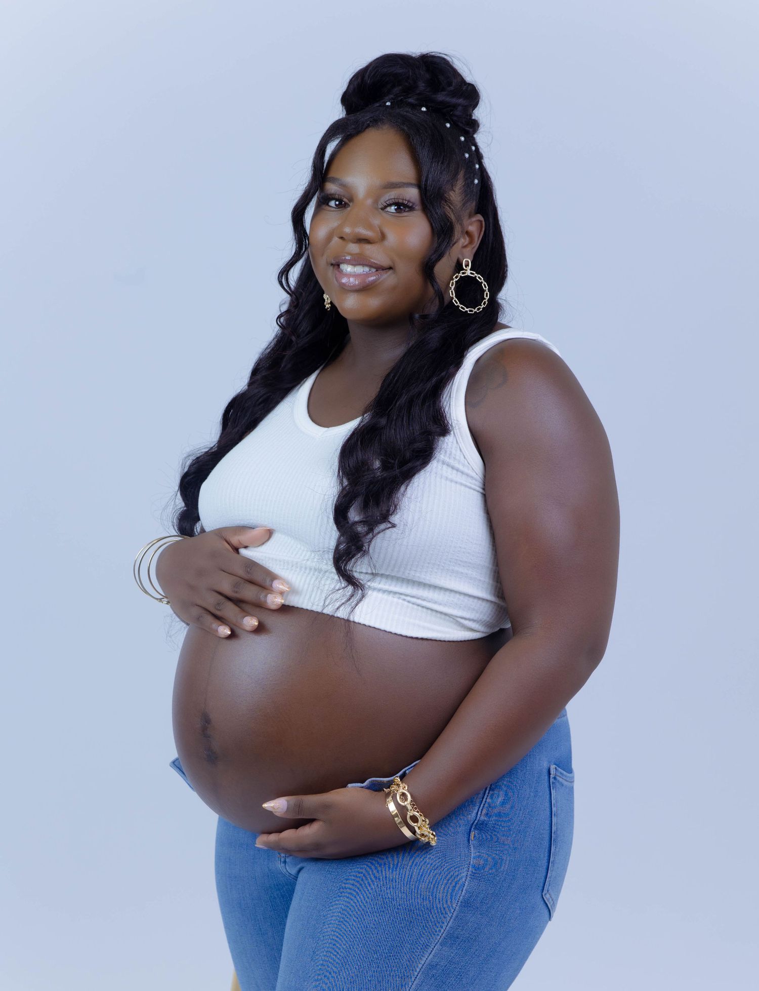 Glowing pregnant woman in white crop top and jeans cradles her baby bump in maternity portrait.