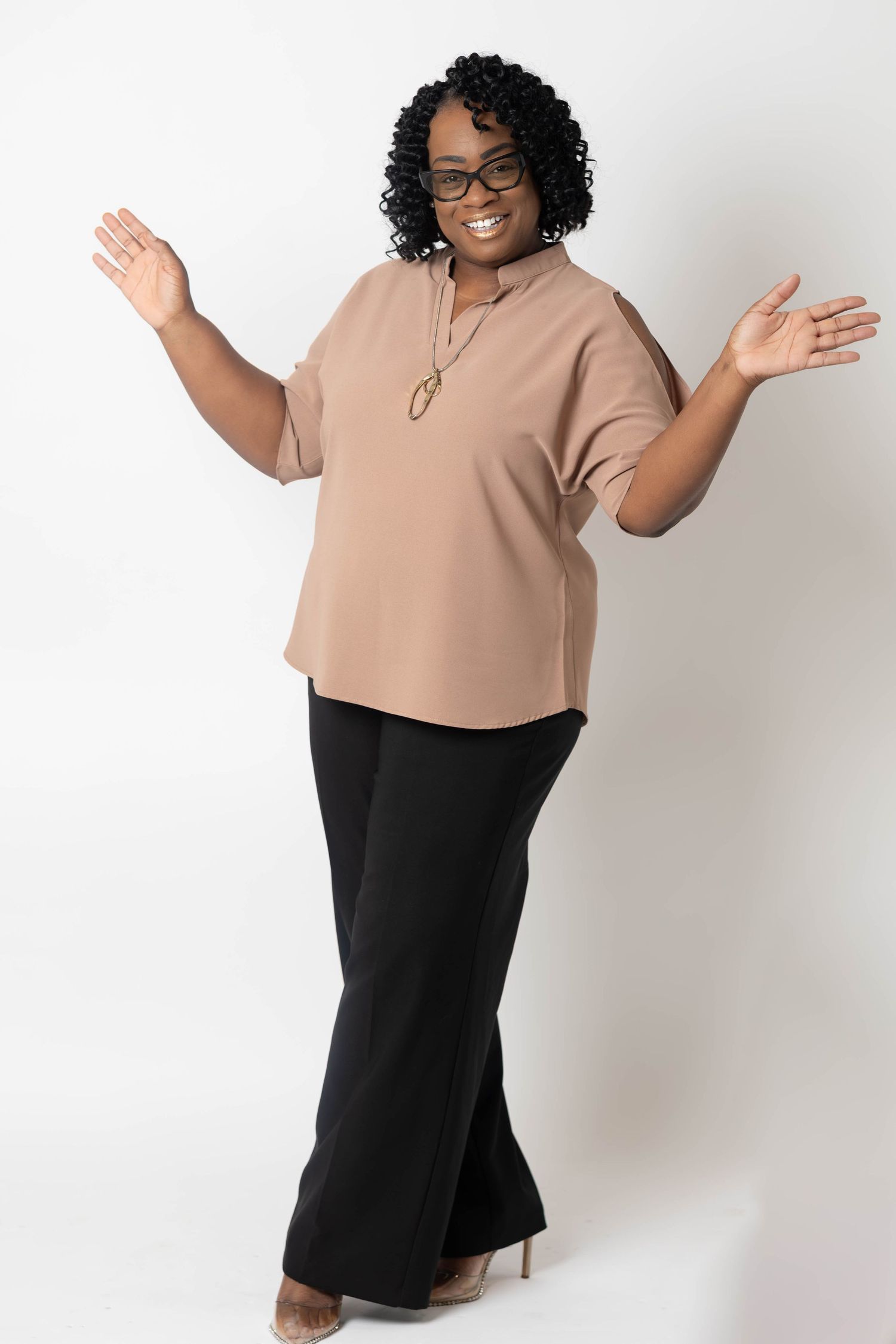 Cheerful plus-size woman in tan top and black pants posing with raised hands on white background.