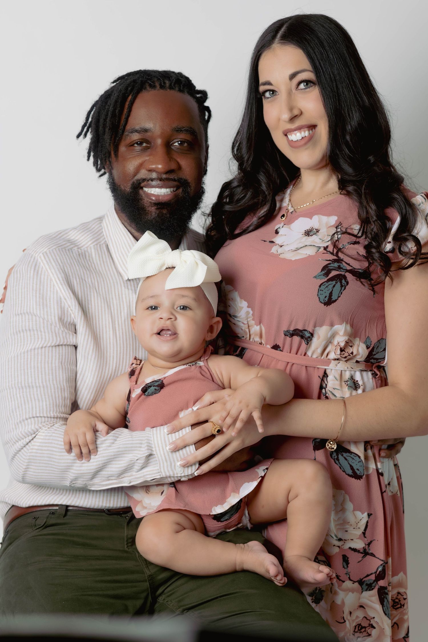 Smiling interracial family portrait with father, mother in floral dress, and adorable baby girl in pink.