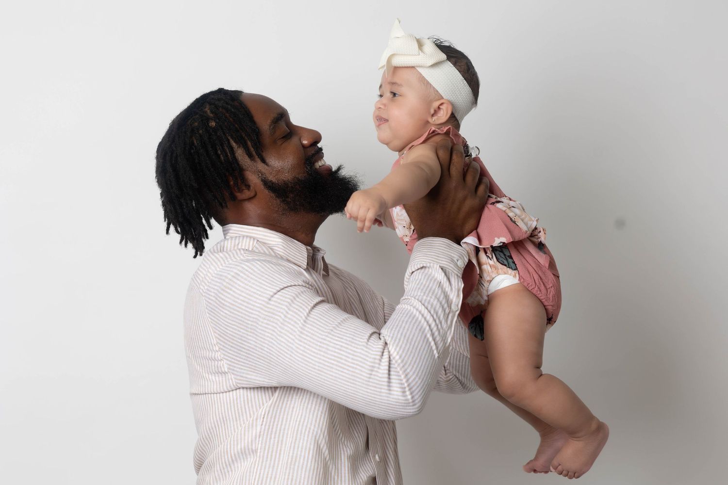 Loving father lifting his smiling baby girl wearing a pink outfit and white bow headband.