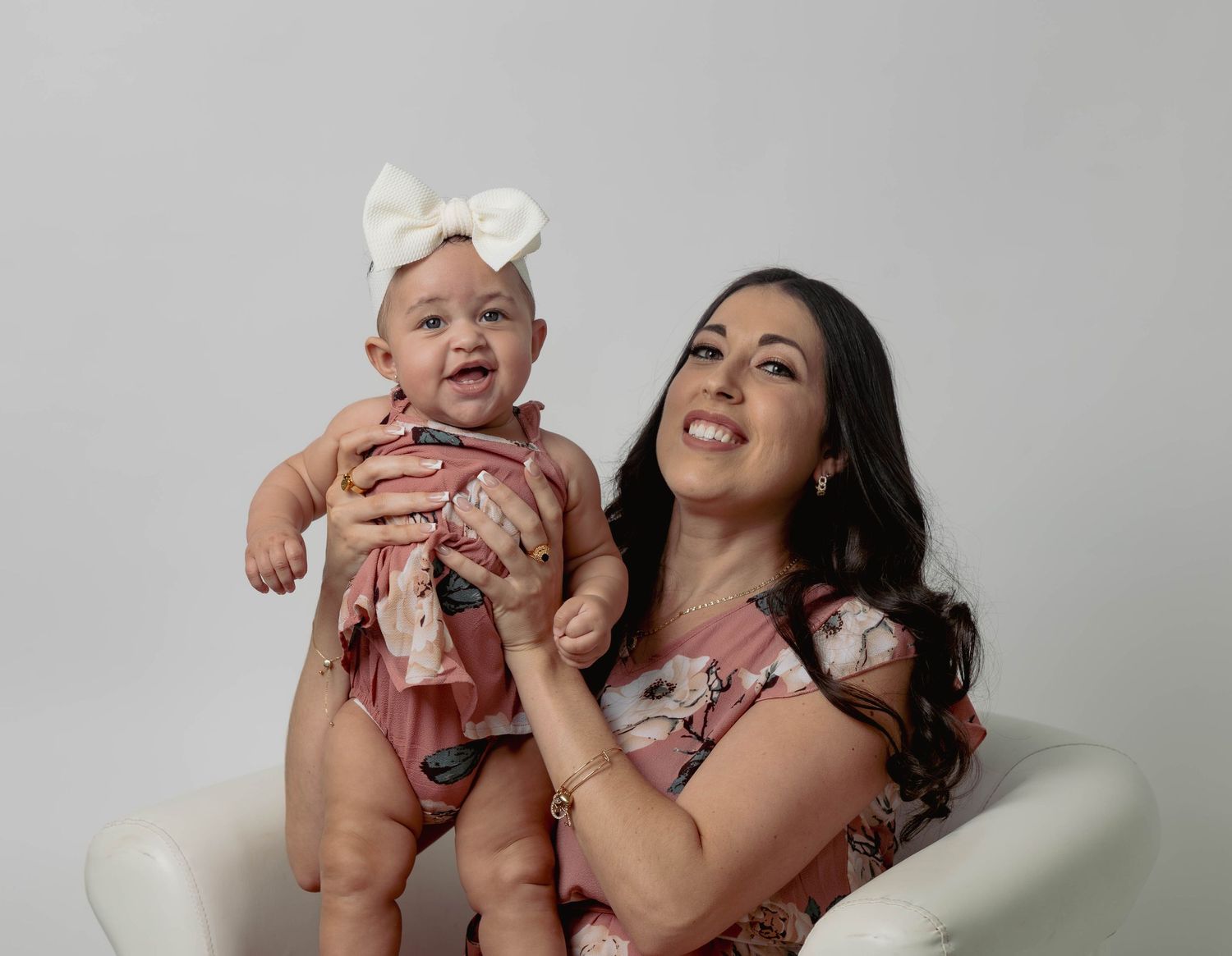 Joyful mother in floral dress holding her baby girl with a large white bow headband on white background.