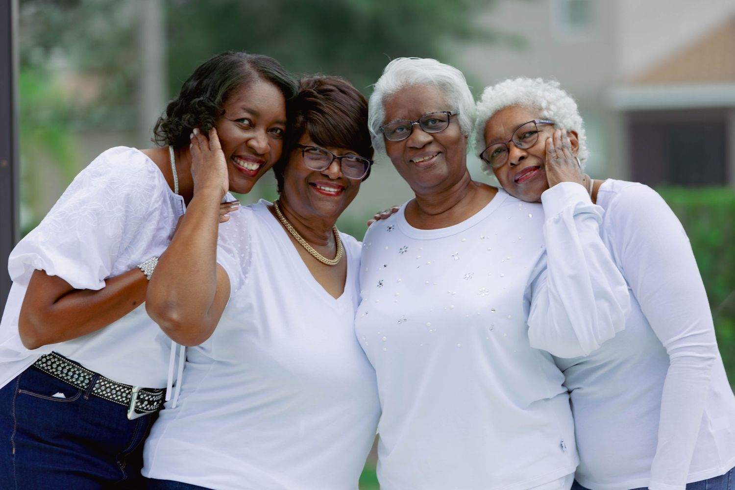 Four generations of Black women smiling together outdoors wearing coordinating white outfits.