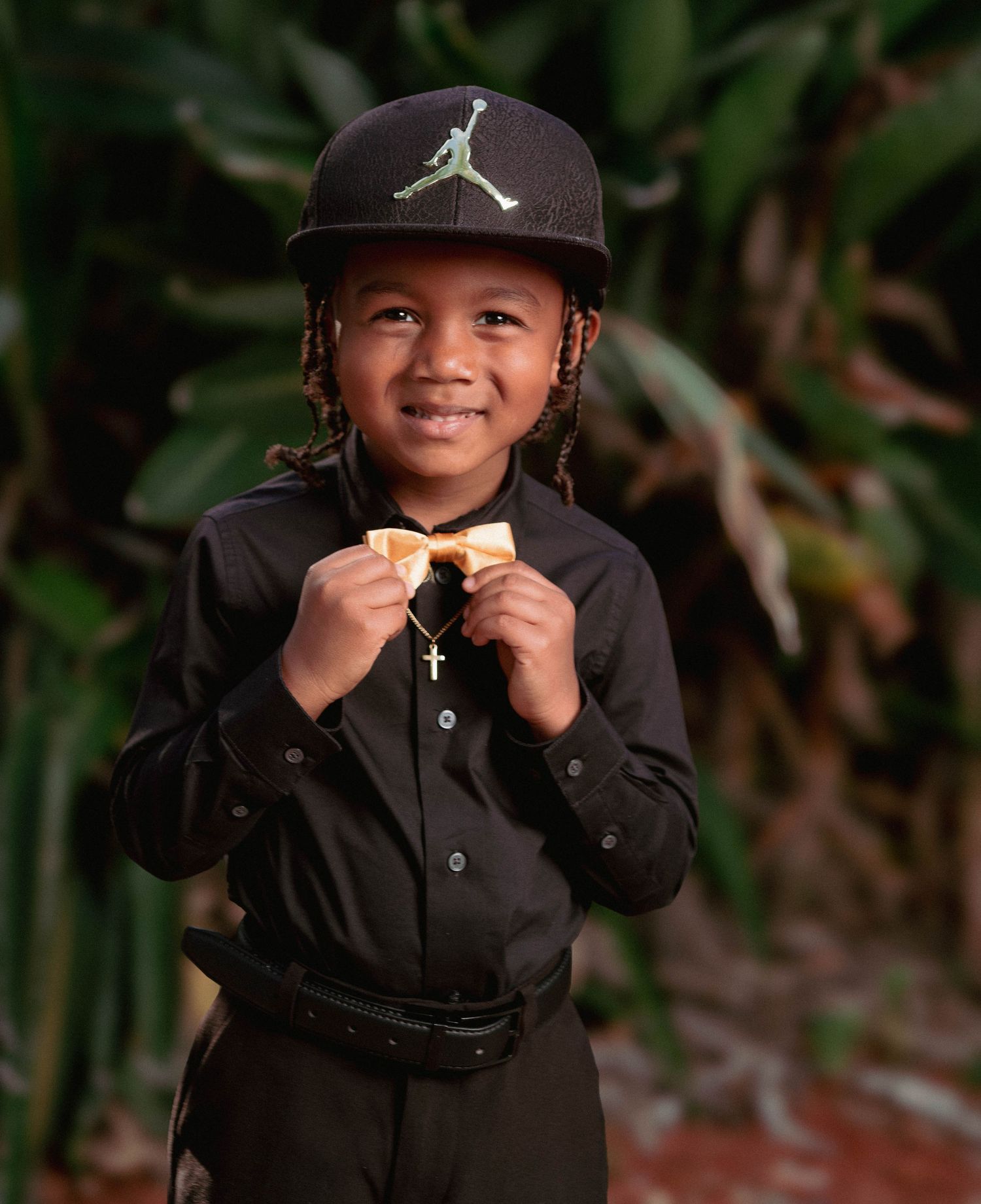 Stylish young boy in black outfit with Jordan cap and gold bow tie smiling in a tropical garden setting.