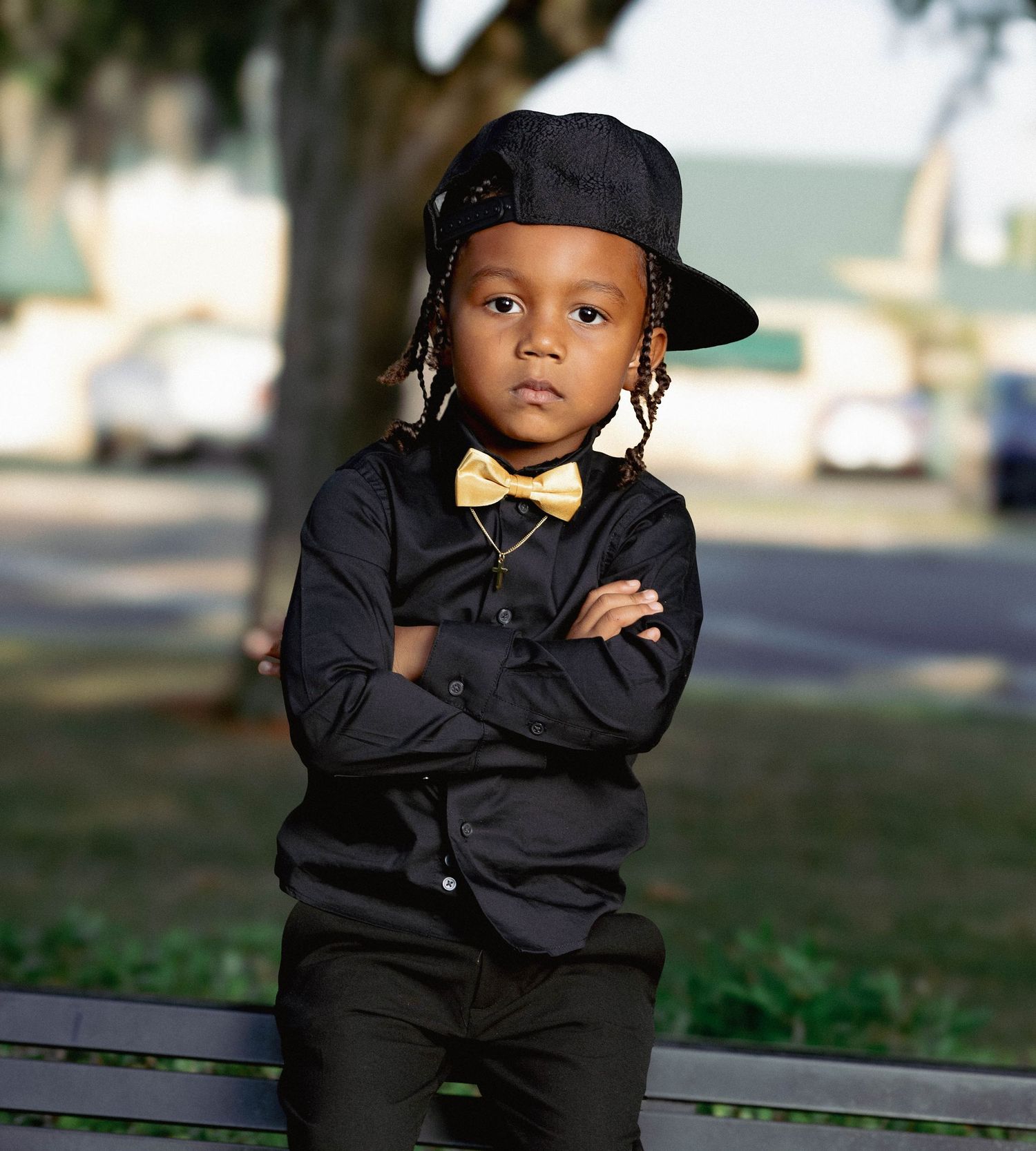 Stylish young Black boy in black outfit with gold bow tie and cap, arms crossed on park bench.