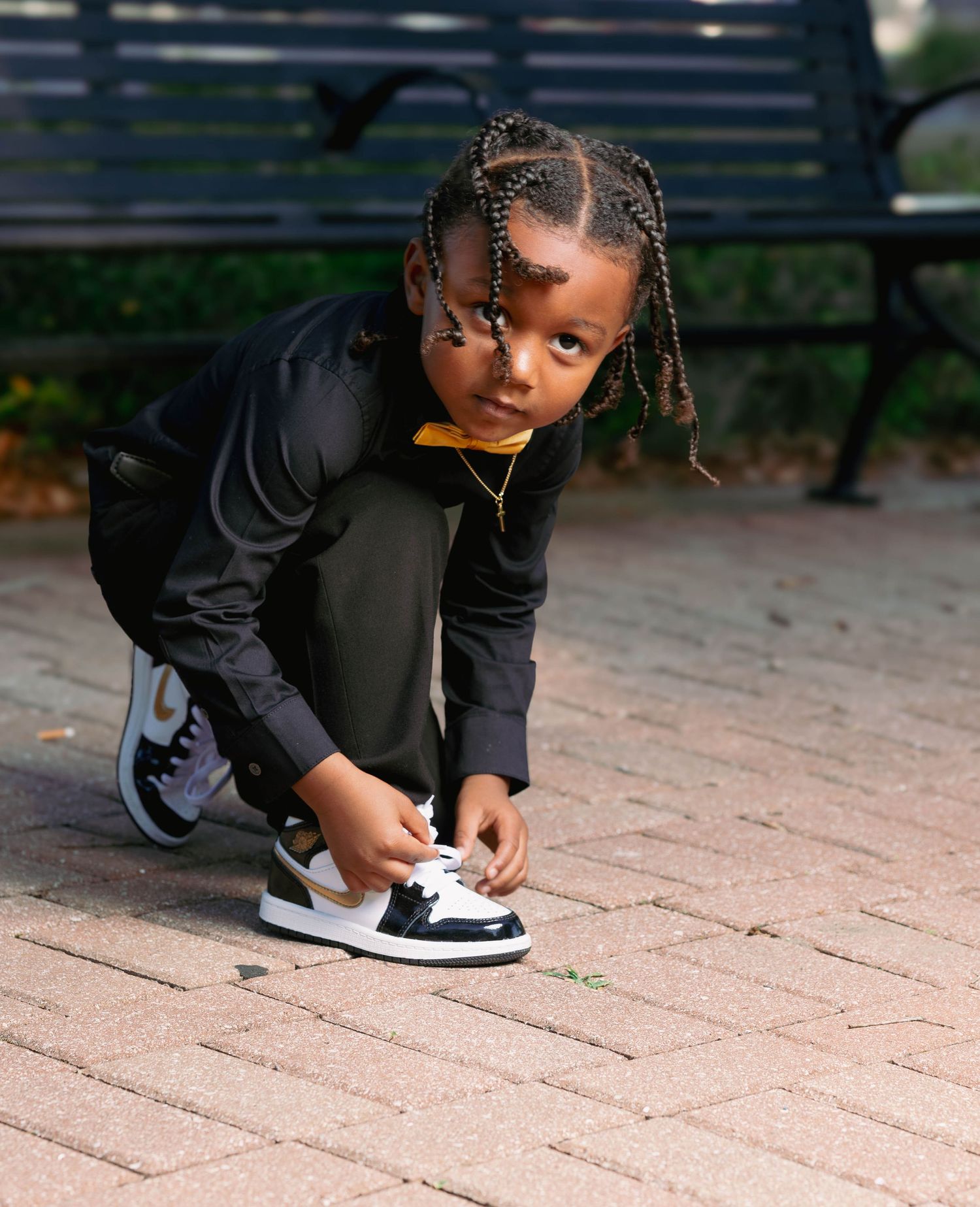 Young Black child with braids tying sneaker laces on brick pavement in a park.
