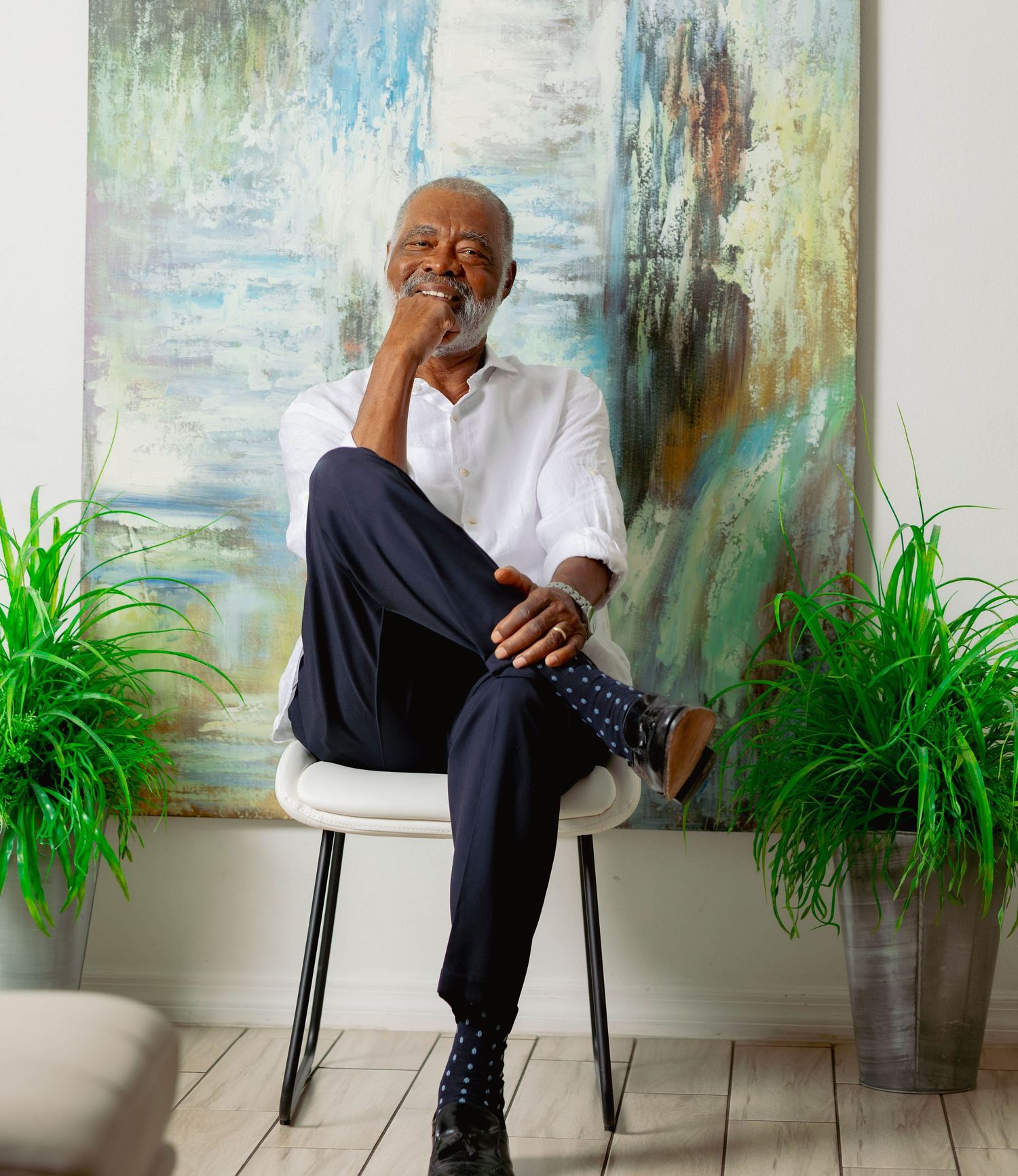 Distinguished older Black man in white shirt seated casually before colorful abstract painting indoors.