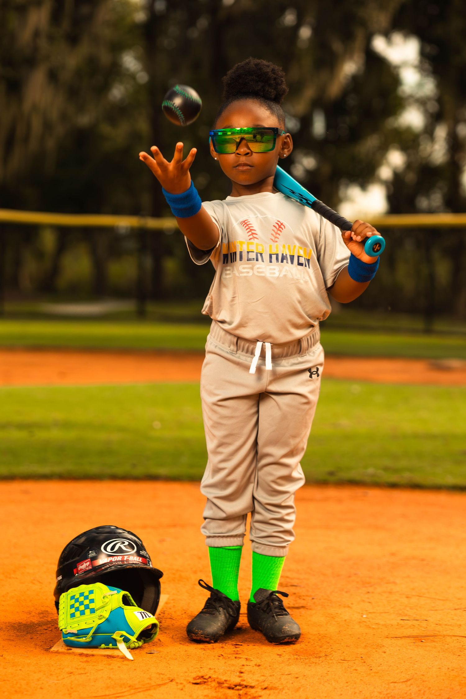 Young baseball player in uniform tosses a ball while holding a bat on a dirt field.
