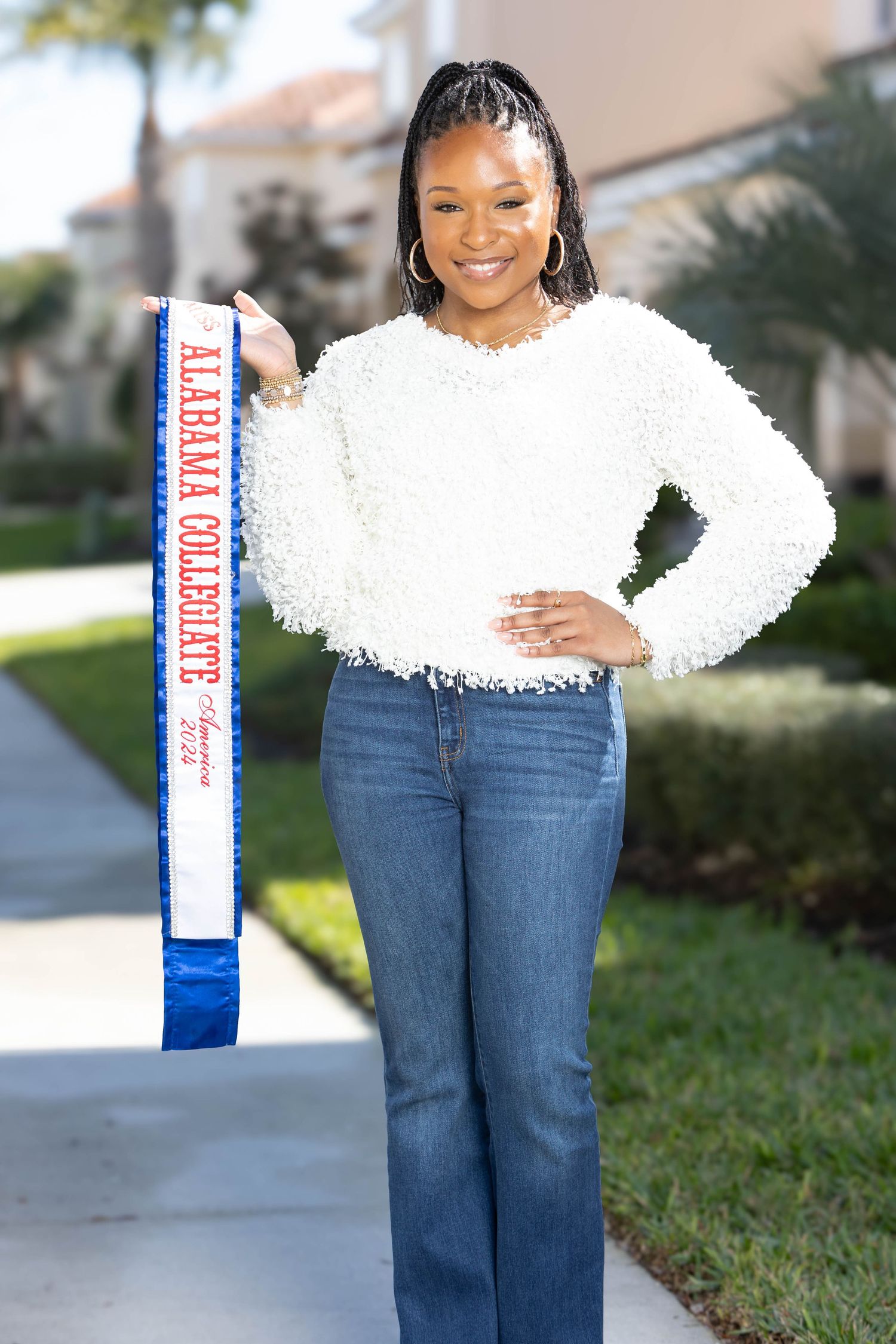 Smiling woman holds Alabama Collegiate sash while posing outdoors in white sweater and jeans.