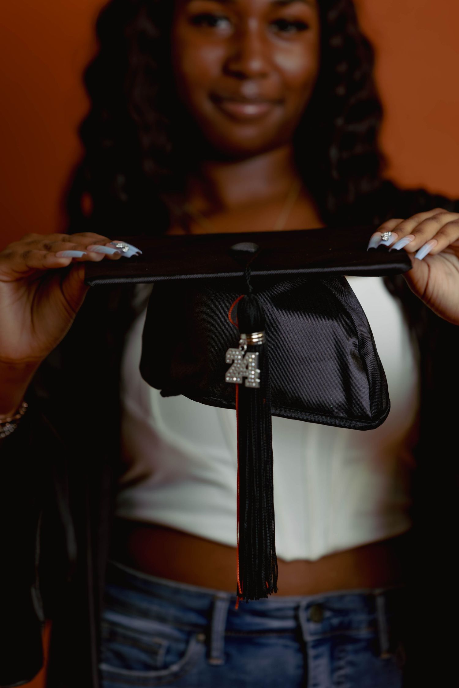 Graduate holding a black cap with Class of 2024 charm against an orange background.
