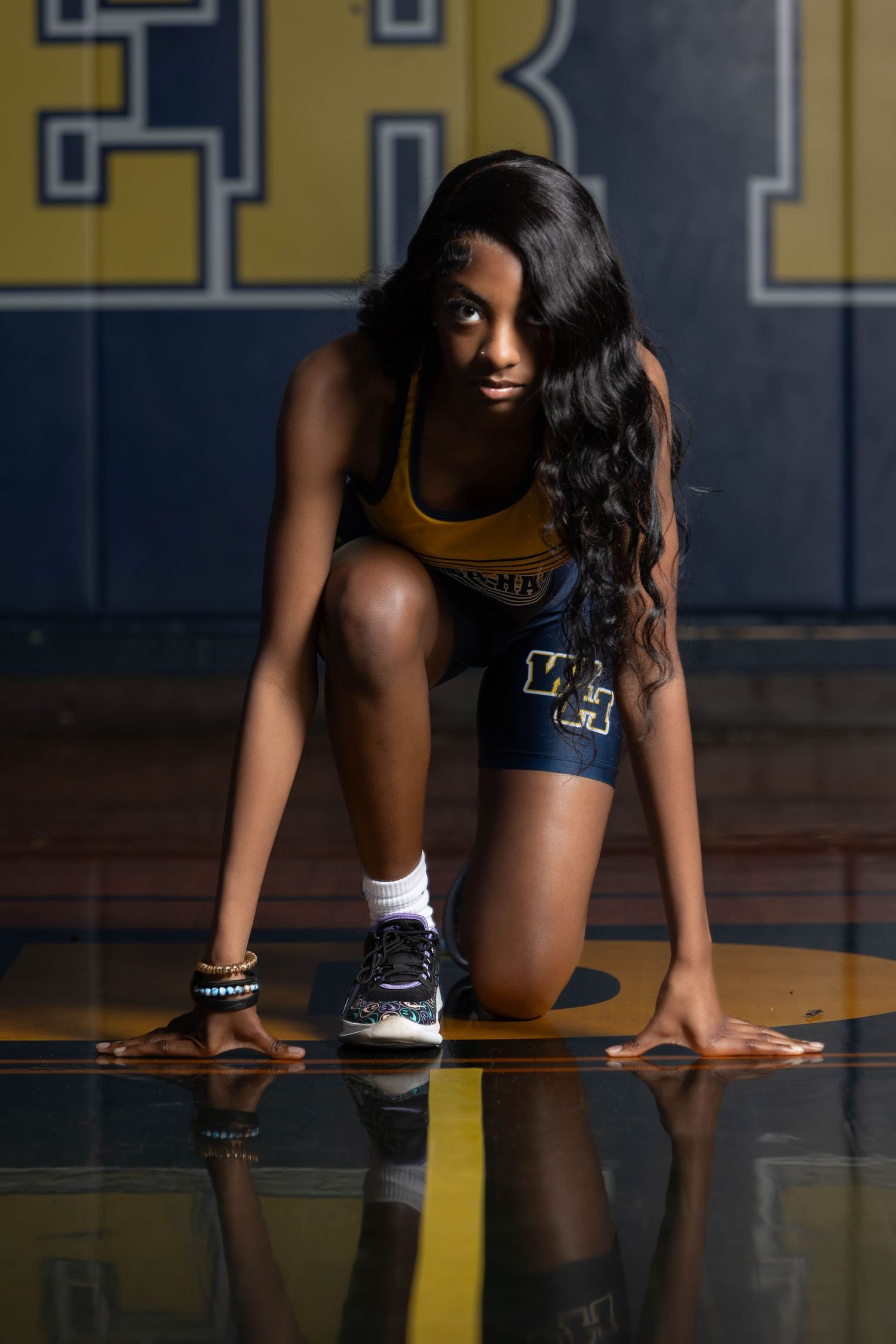 Female athlete in navy and gold uniform crouches in starting position on a gymnasium floor.