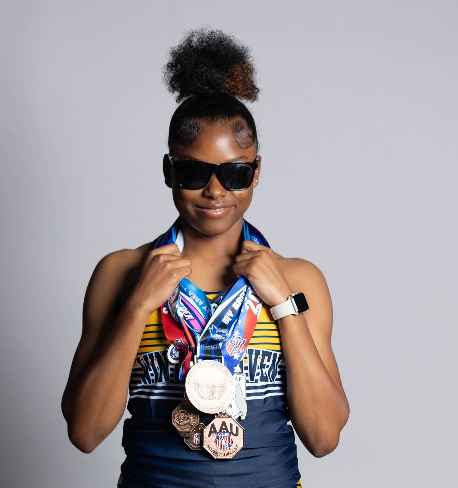 Young female athlete proudly displays multiple AAU medals while wearing sunglasses and athletic wear.