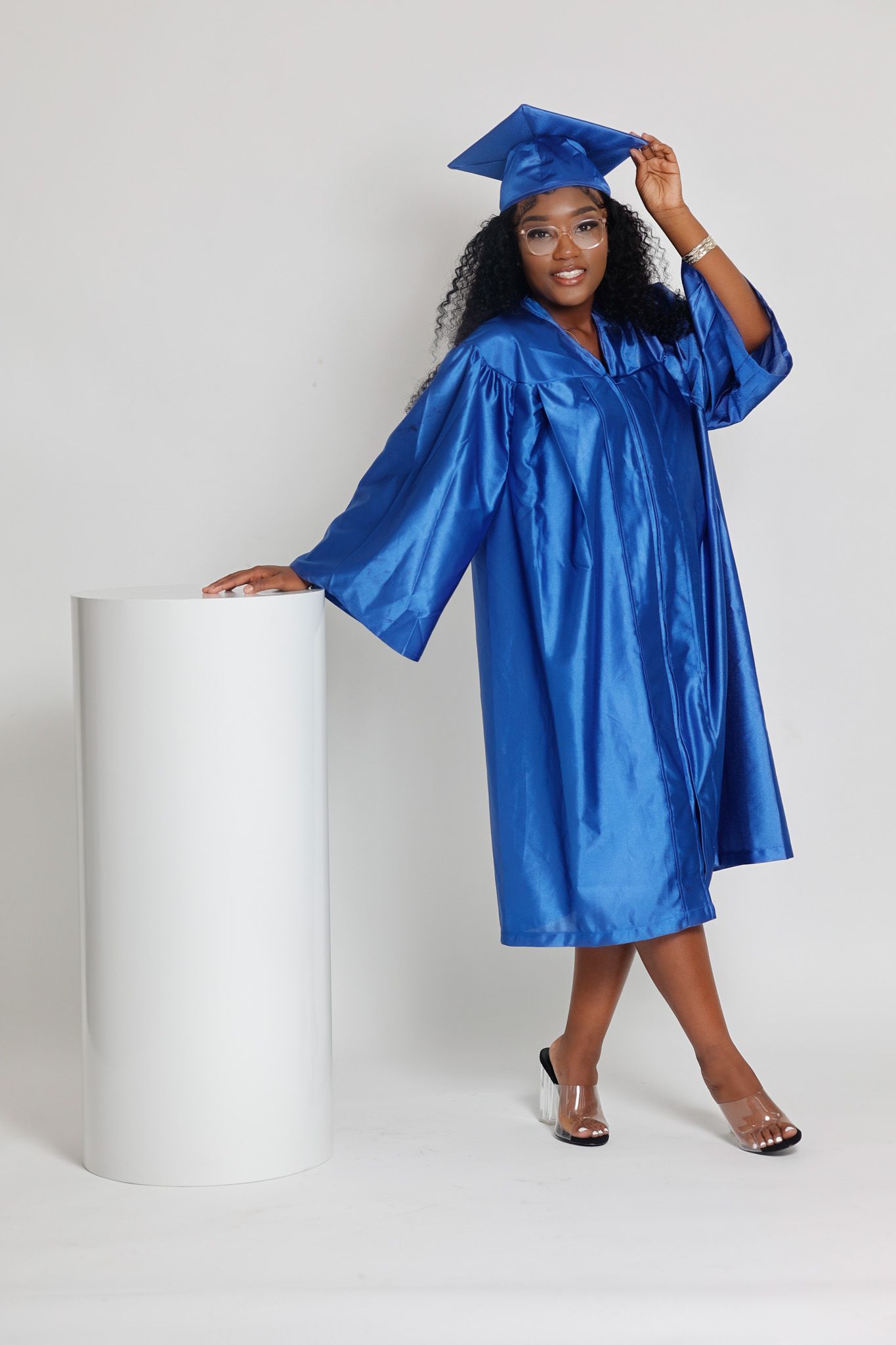 Young Black woman in royal blue graduation cap and gown posing beside white cylinder prop.