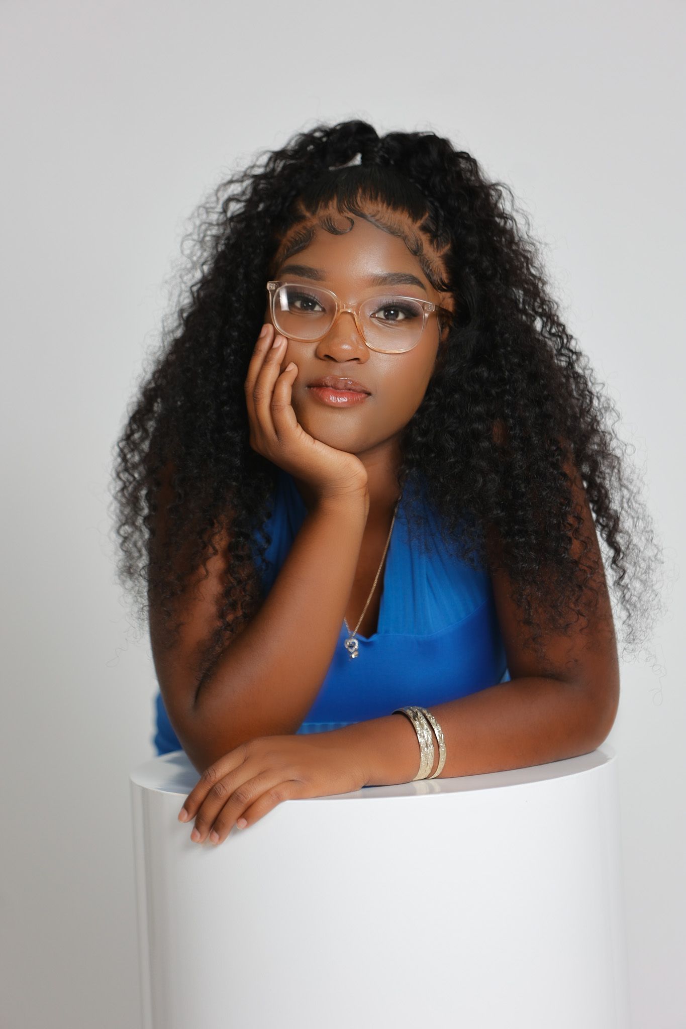 Young Black woman with curly hair and glasses leaning on white cylinder prop in blue top.