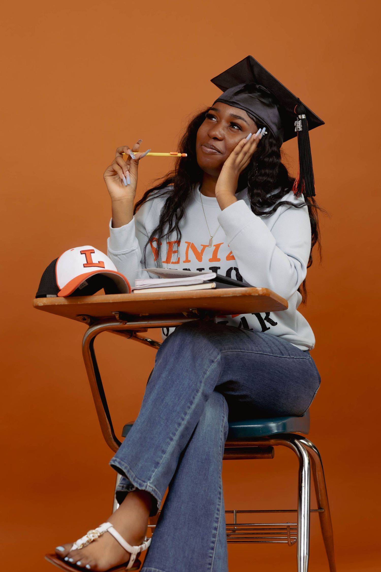 Graduate sitting at school desk wearing cap and university sweatshirt in a thoughtful pose.