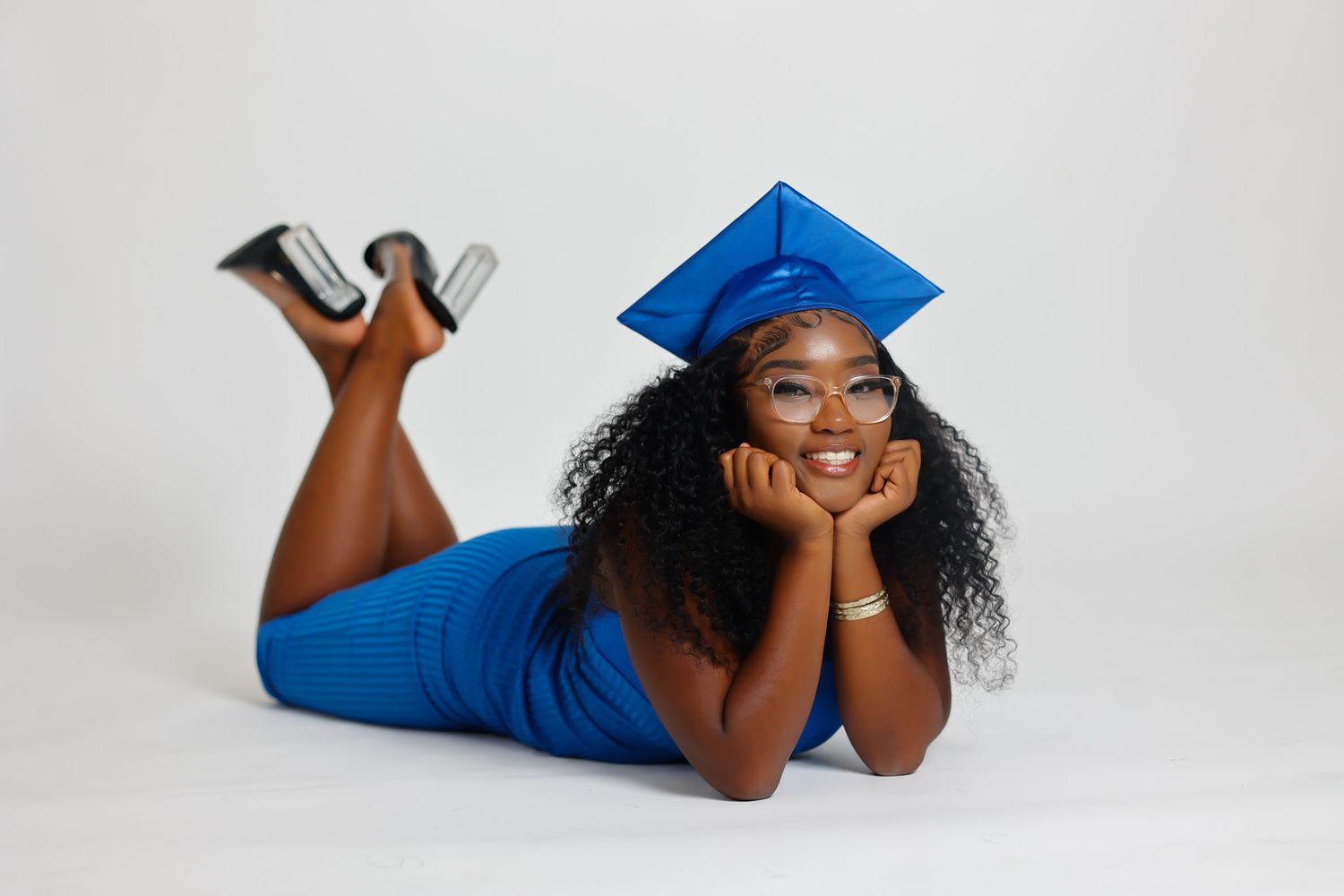Young Black woman in blue dress and graduation cap lying down smiling holding diploma.