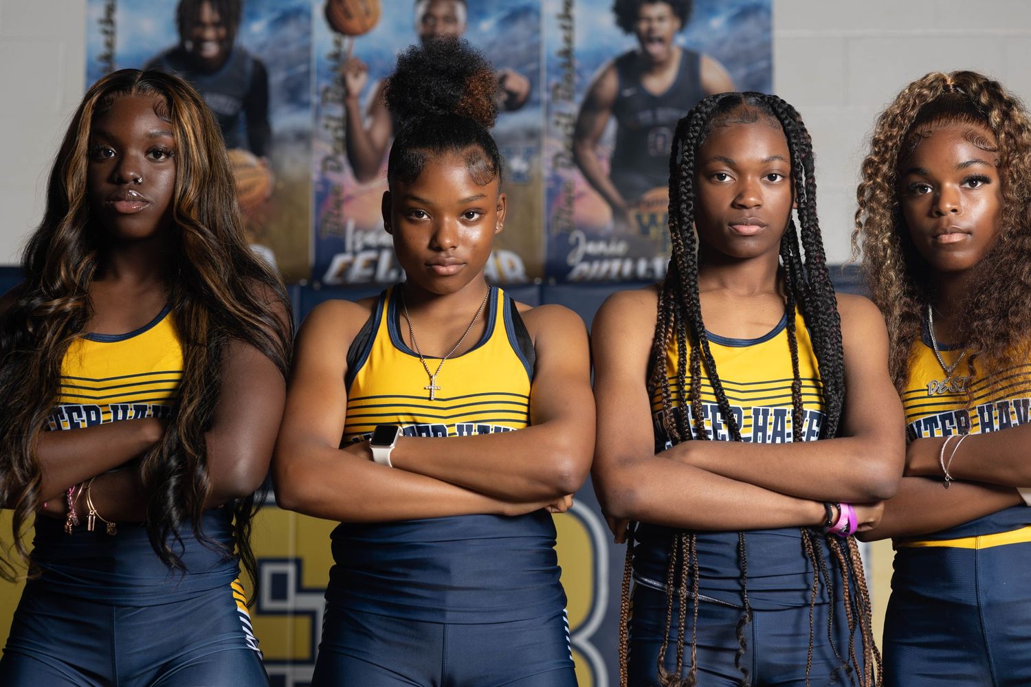 Four young female athletes in navy and gold uniforms pose with arms crossed in a gymnasium.