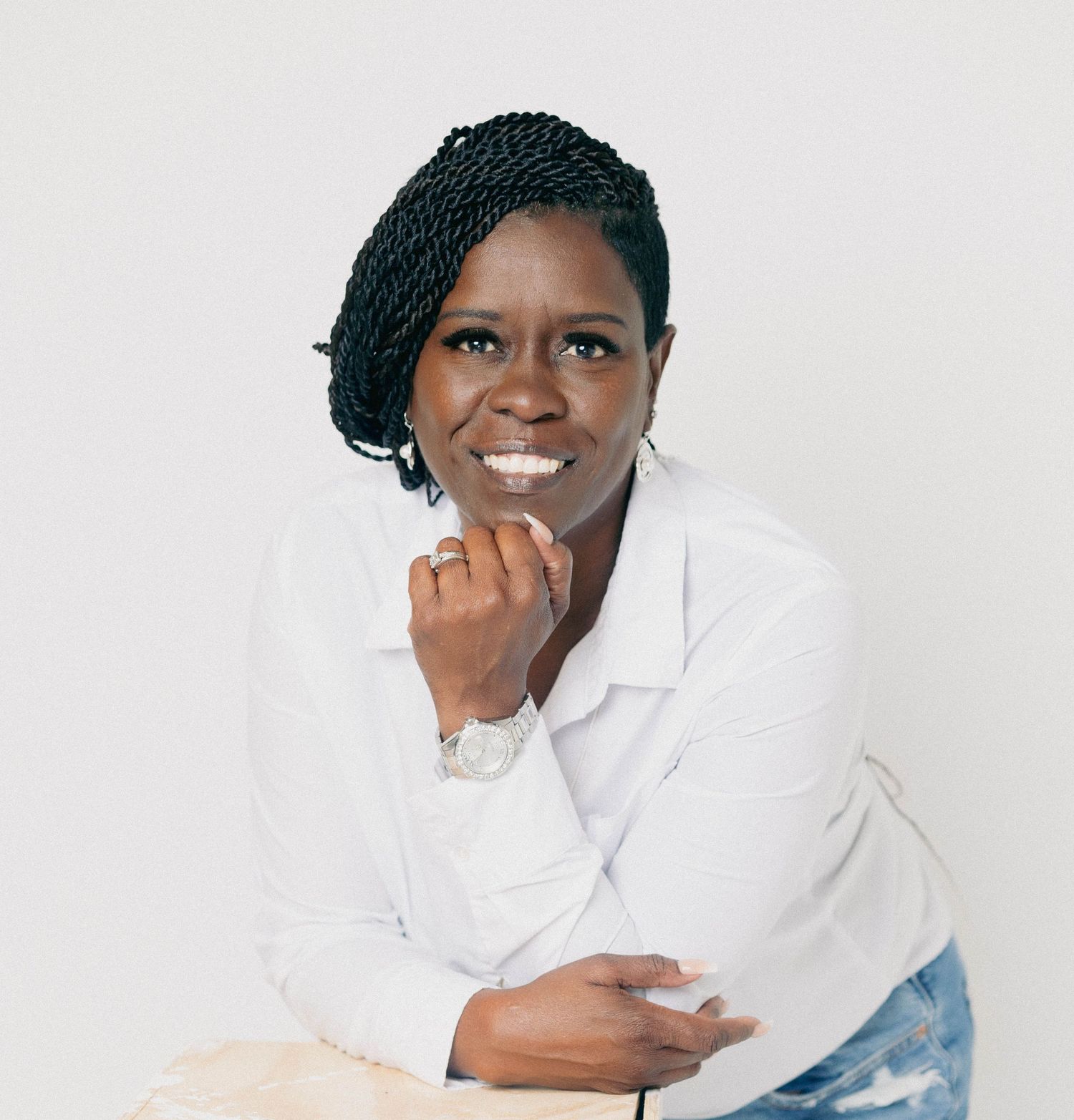 Smiling woman in white blazer poses with hand near chin in bright studio setting.