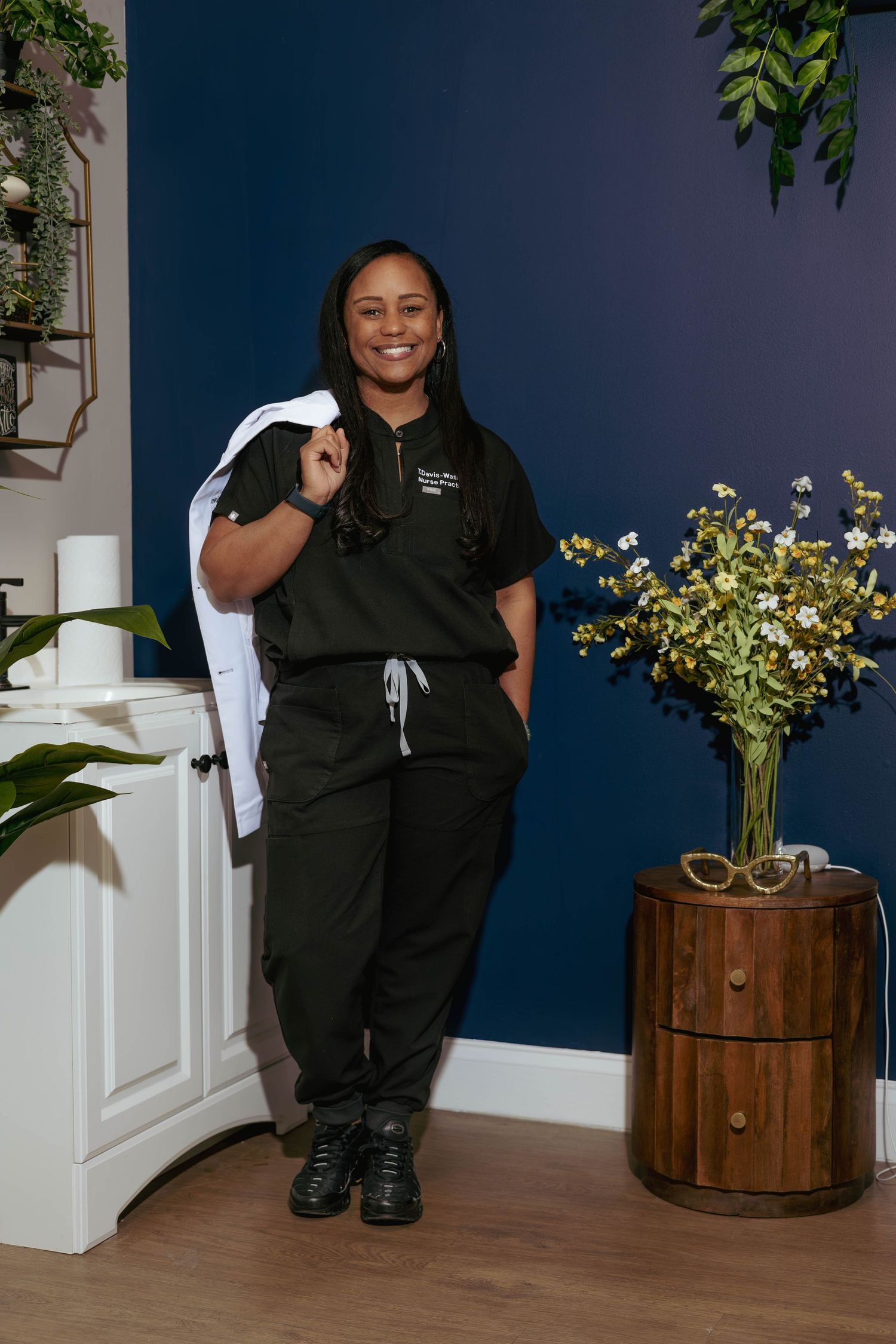 Black female medical professional in dark scrubs smiling in a modern clinic interior.