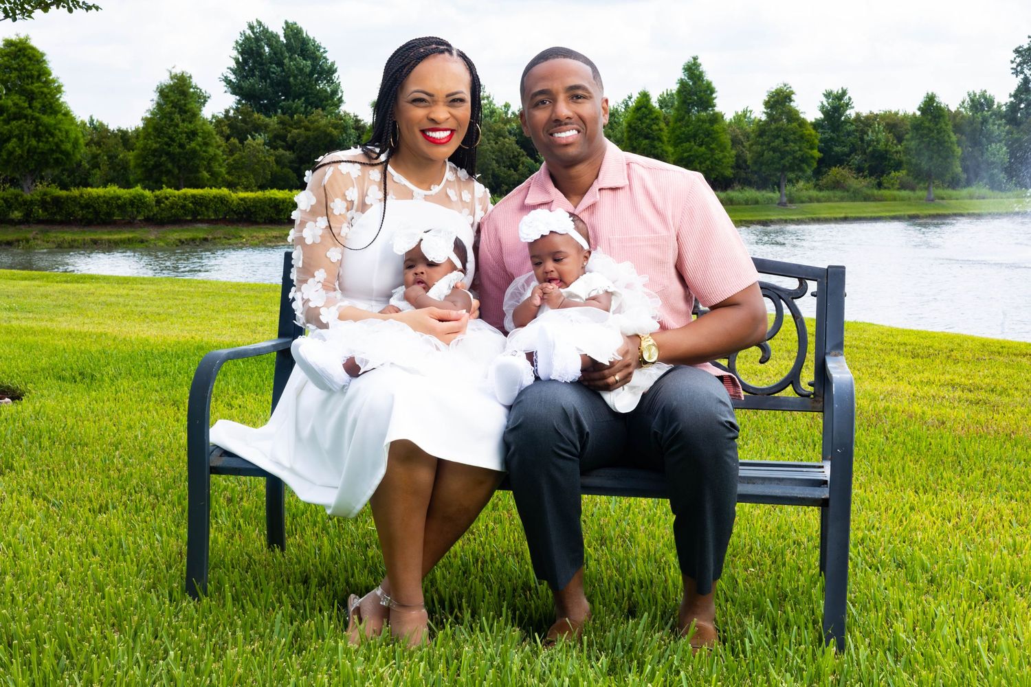 Happy family portrait on park bench with parents holding twin babies in white outfits near a scenic pond.
