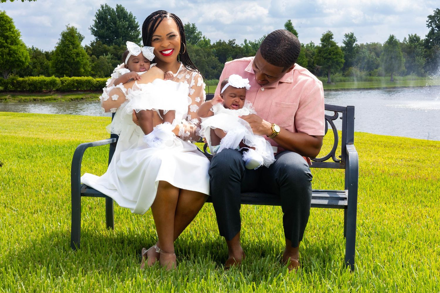 Couple sitting on a park bench holding twin babies in white outfits with a serene pond in the background.