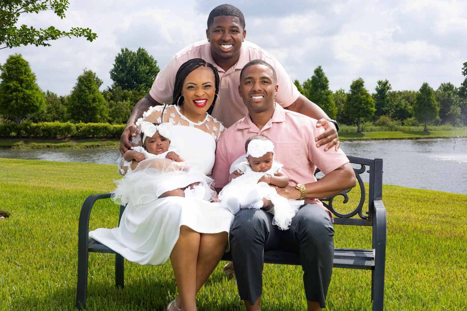 Happy family of four posing on a bench outdoors, parents holding twin babies in white dresses near a pond.