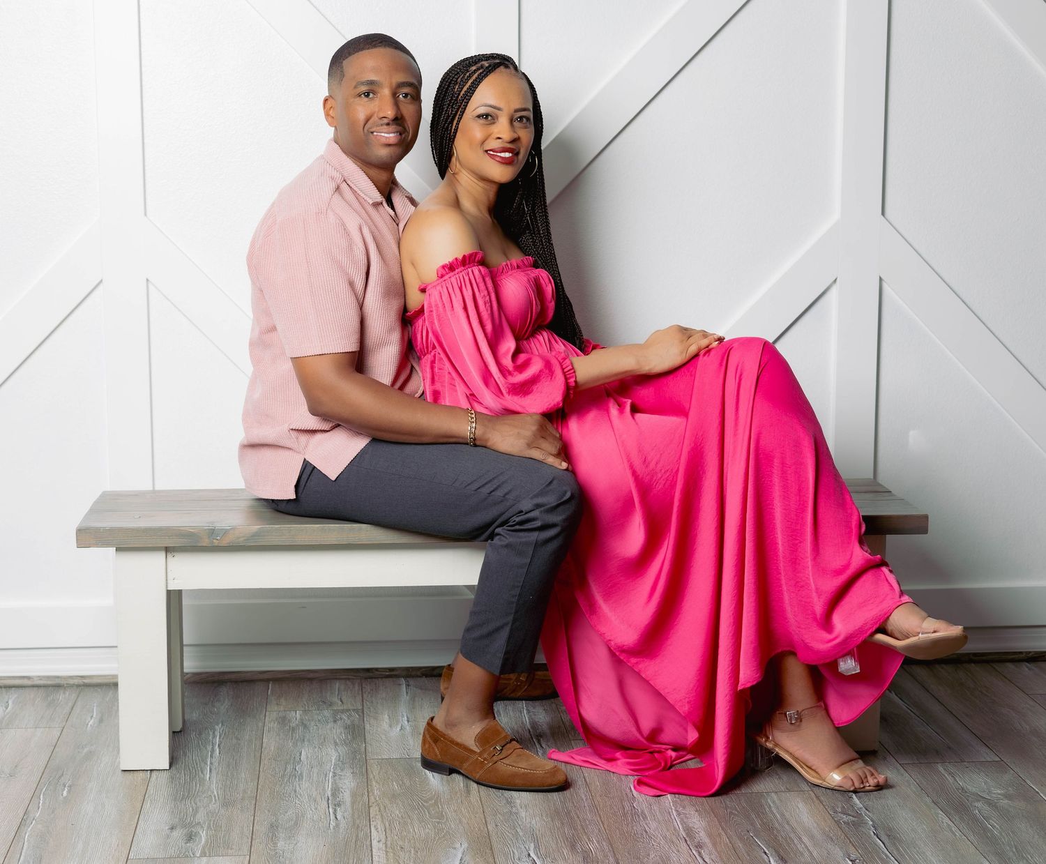 Elegant couple posing on bench, man in pink shirt and woman in flowing hot pink off-shoulder gown.