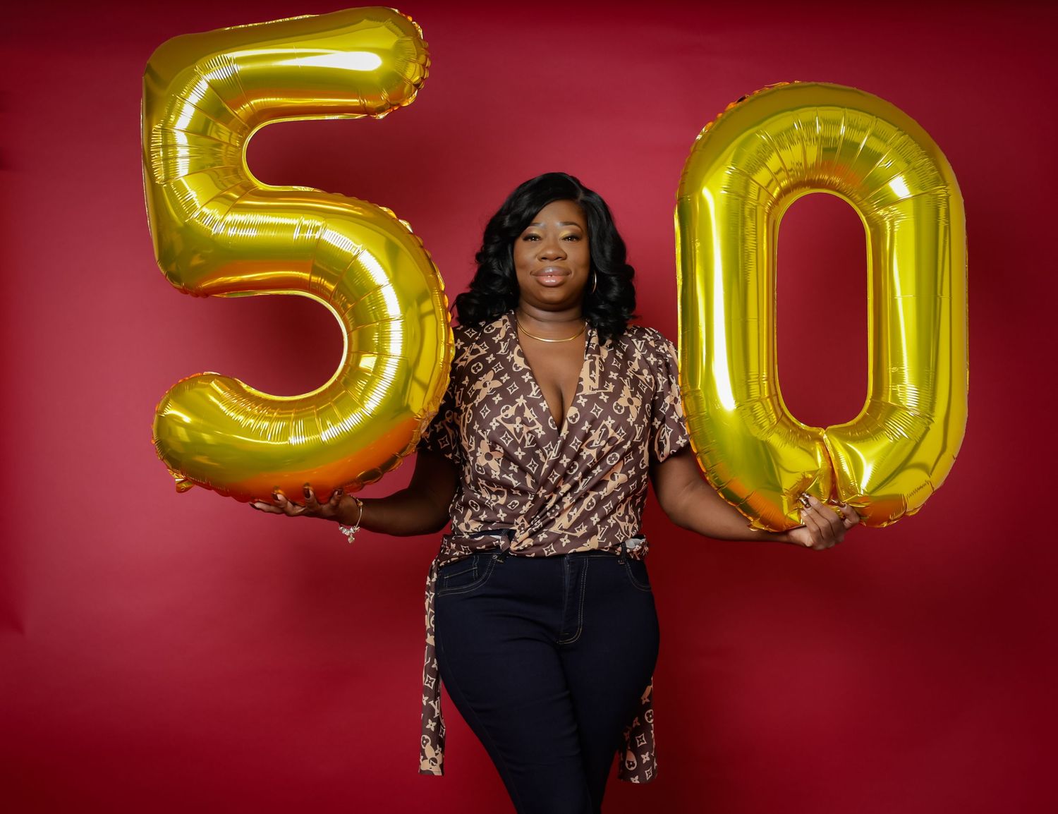 Woman holding gold number 50 balloons wearing patterned blouse against vibrant red background.