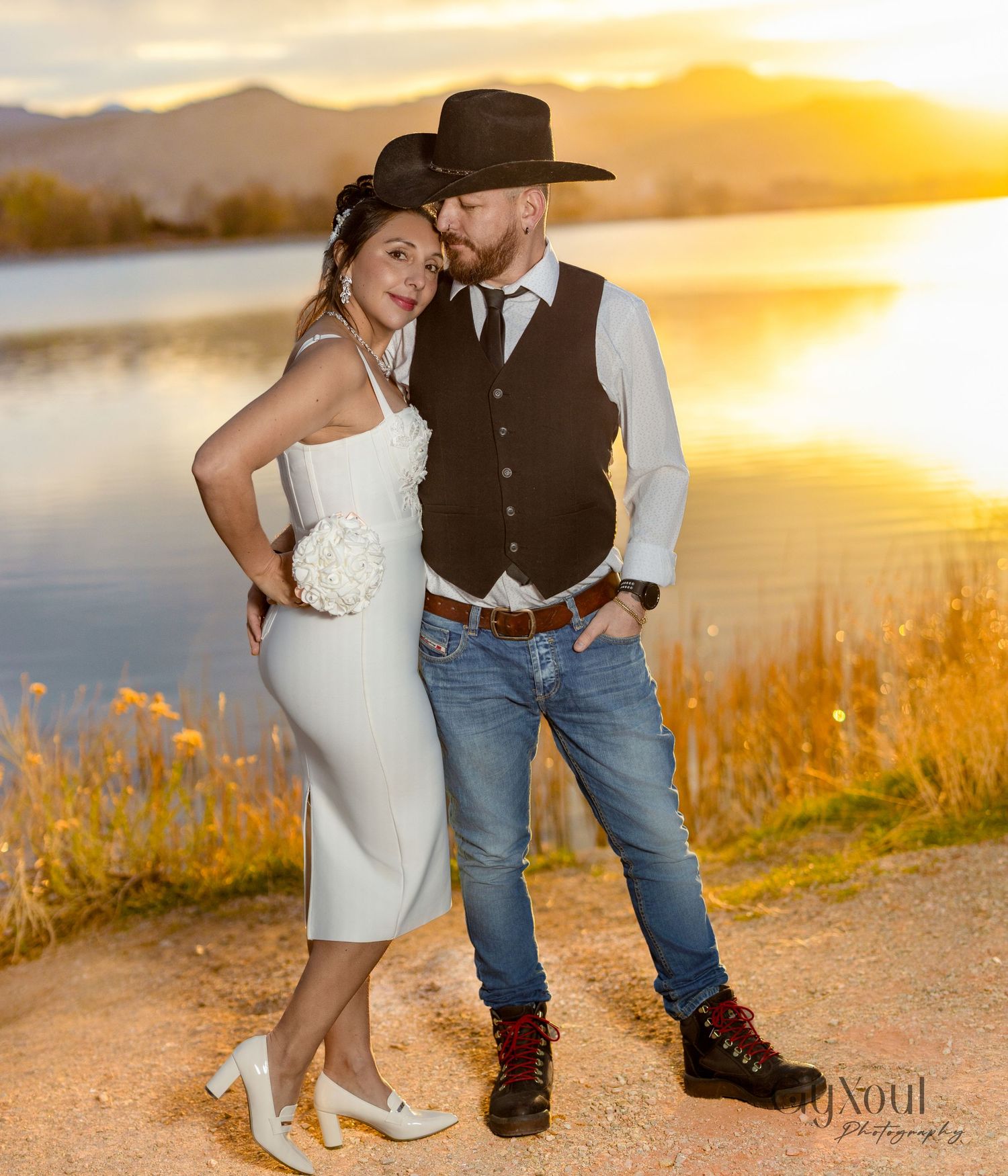 A couple poses together at sunset by a lake, with golden light reflecting off the water and mountains in the background.