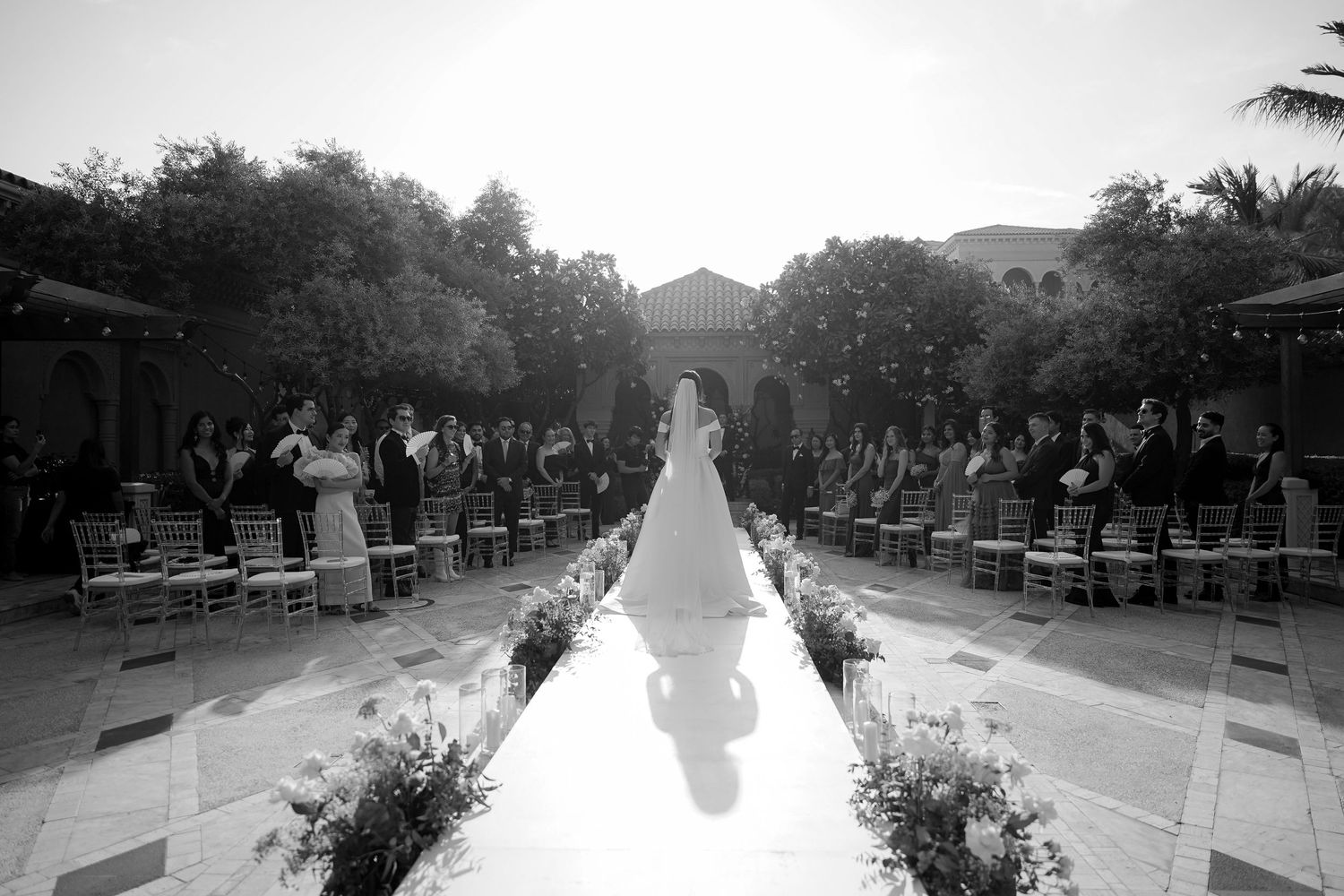 Black and white image of an outdoor wedding ceremony with a white aisle runner and floral decorations between rows of chairs.