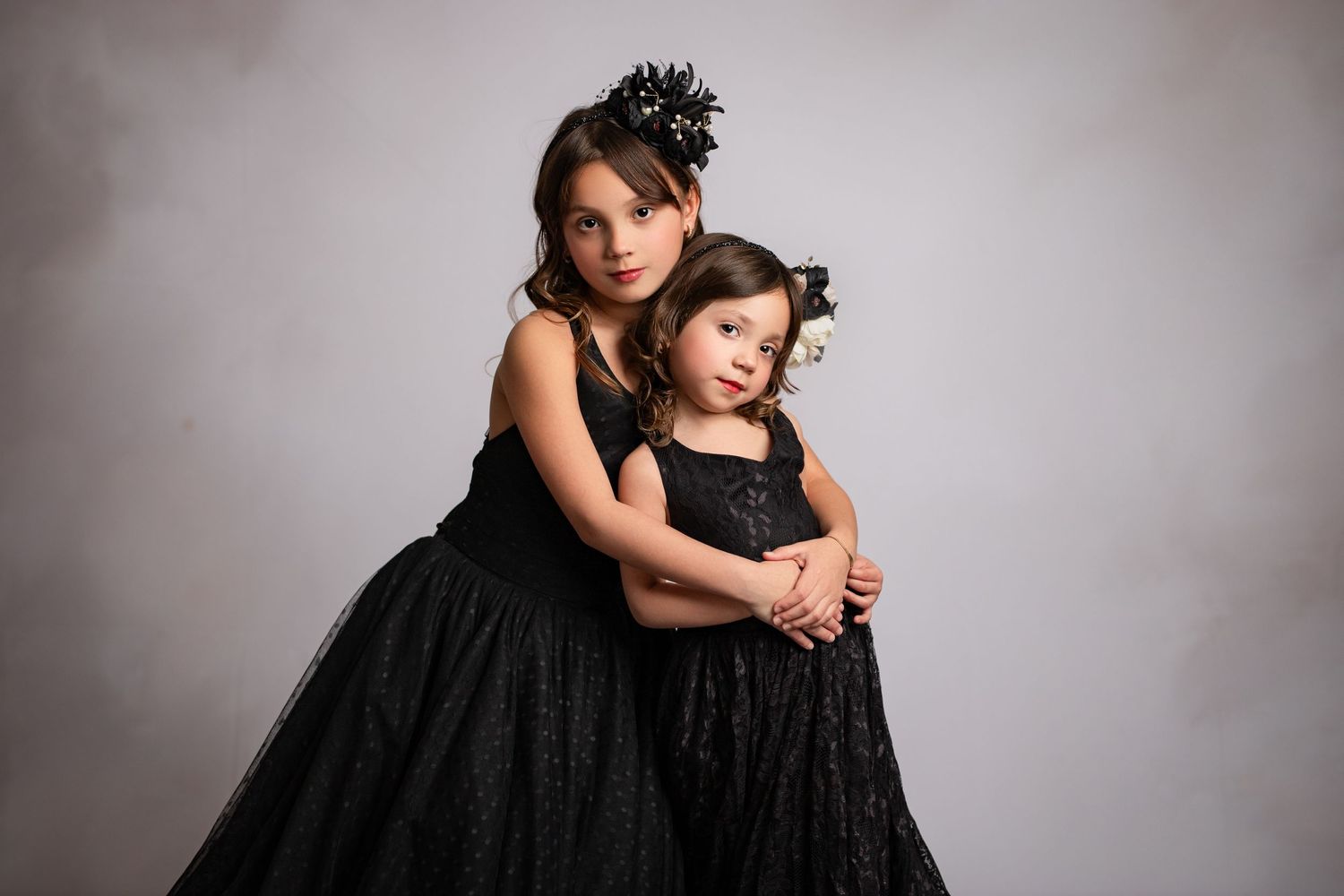 Two people in matching black tulle dresses pose together against a gray studio backdrop for a formal portrait.
