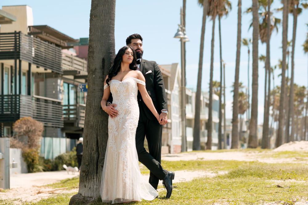 A couple in wedding attire poses together against a backdrop of palm trees and modern beach houses on a sunny day.