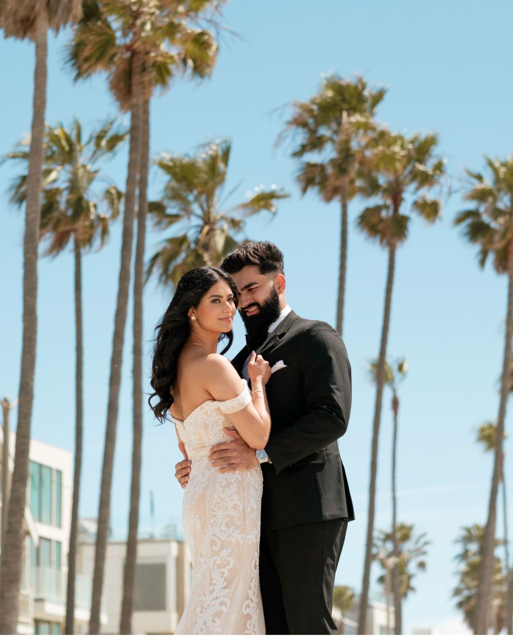 A couple shares a romantic moment on a sunny day with tall palm trees swaying in the background.
