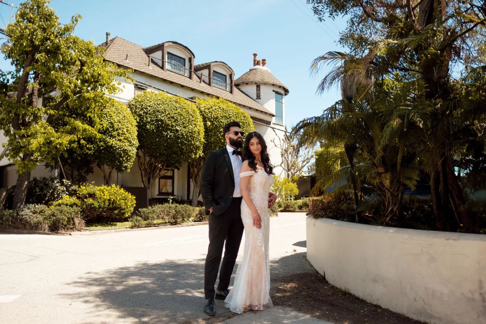 A couple stands together in front of a beautiful Cape Cod style house with lush landscaping on a bright sunny day.