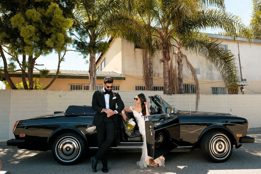 A couple in formal attire sits by a vintage black Rolls Royce on a sunny day with palm trees in the background.