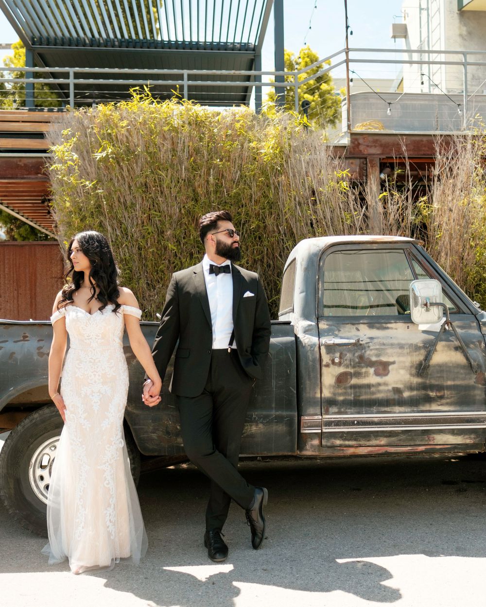 A couple in wedding attire poses beside a vintage black pickup truck with yellow flowers in the background.