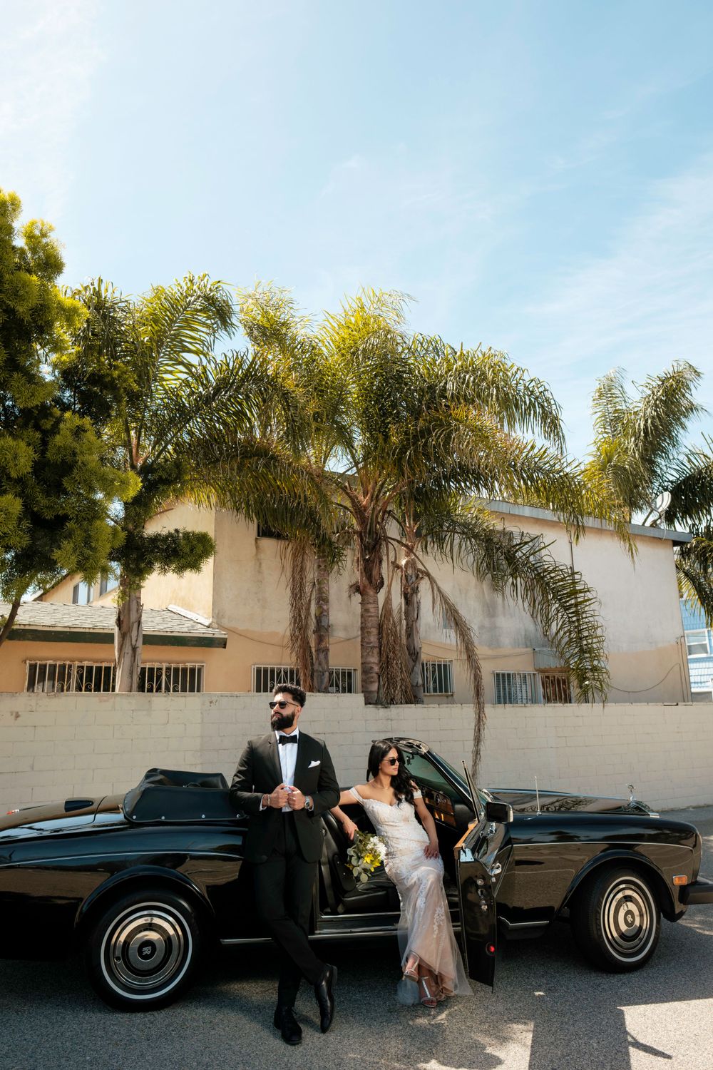 A couple poses with their classic black convertible car in front of palm trees on their wedding day.