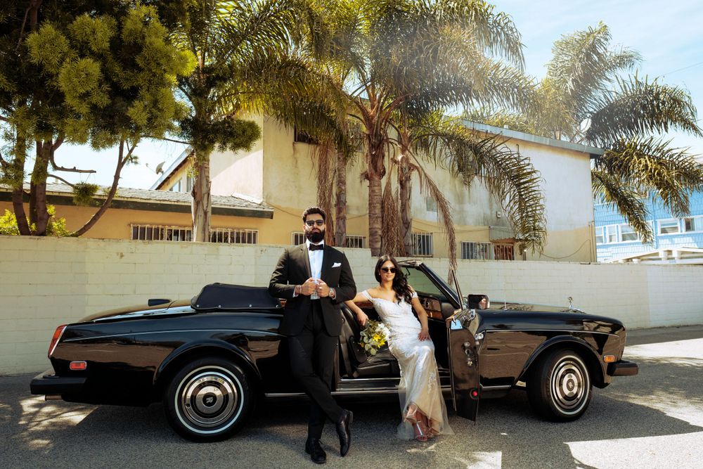 A couple poses beside a classic black car on a sunny day with palm trees and a white building in the background.