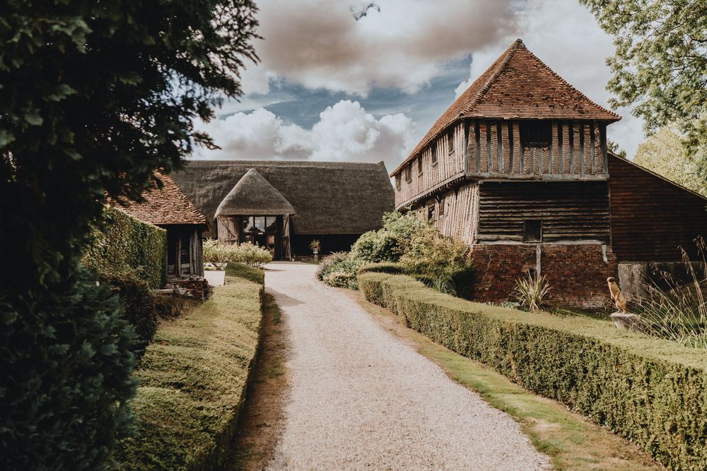 A winding gravel path leads through rustic medieval barn buildings  of Colville Hall at an English heritage site on a cloudy day.