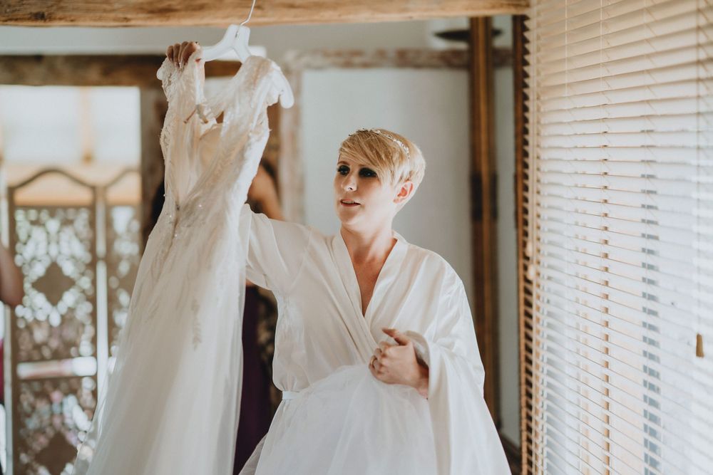An elegant white wedding dress hangs from a hanger as someone in a white robe examines it by a window.