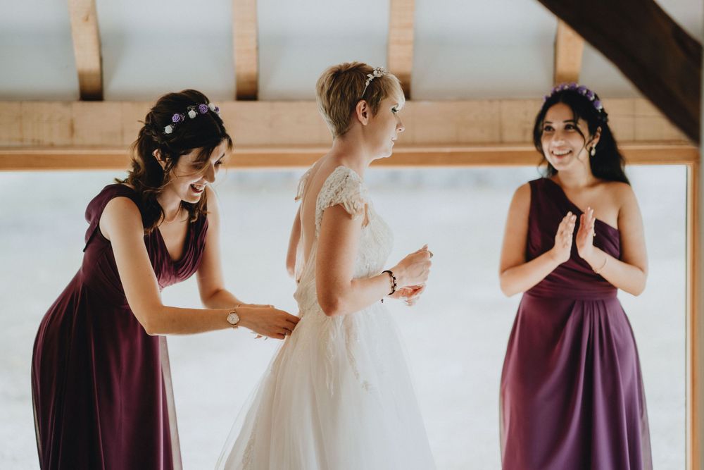 Bridesmaids in burgundy dresses help adjust the wedding gown in a light-filled room during preparations.