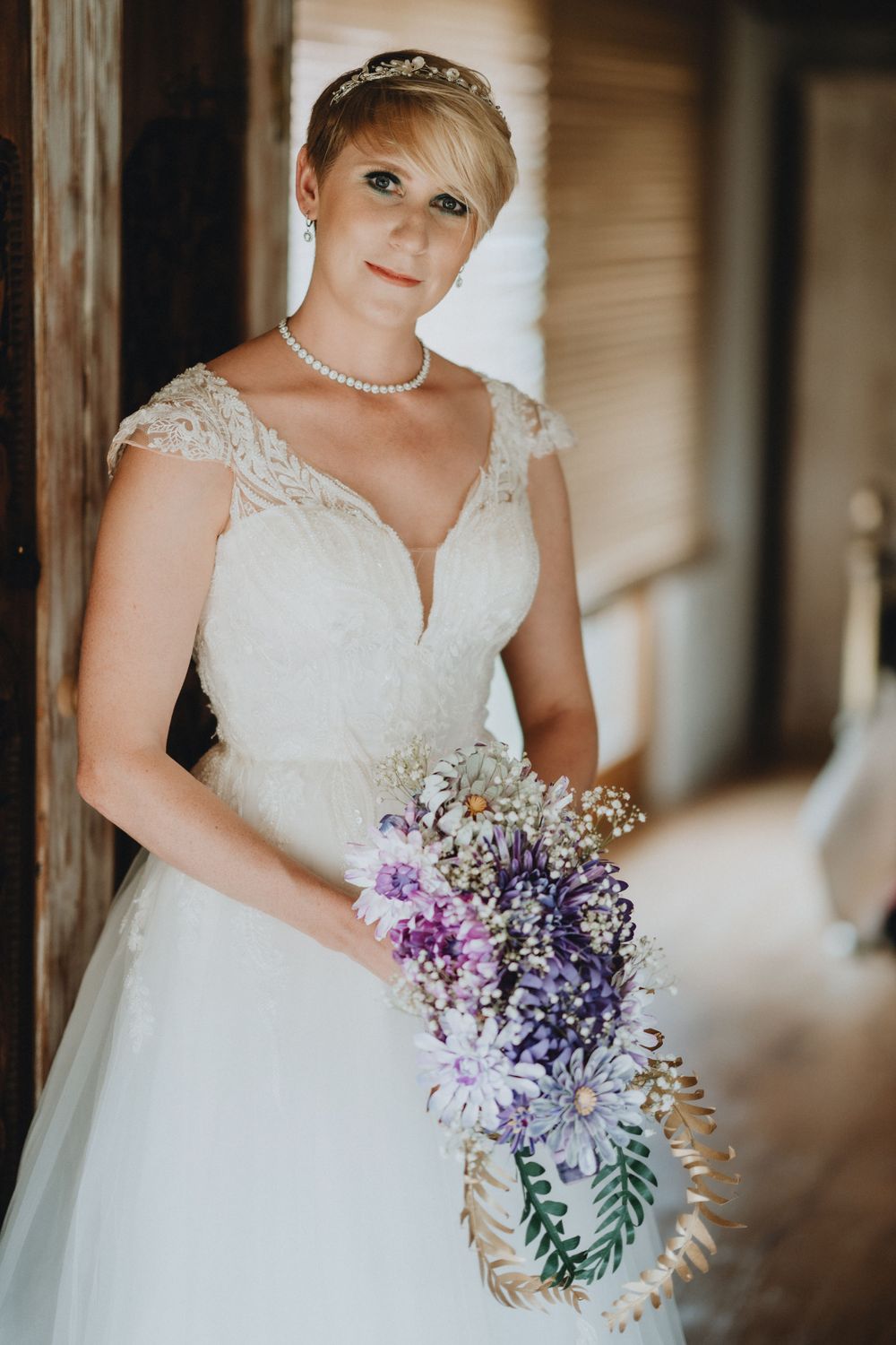 Bride in flowing white lace wedding gown with cap sleeves holds elegant purple and white floral bouquet.