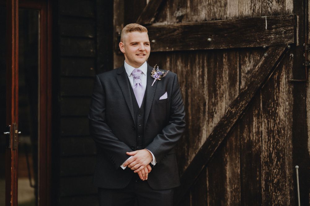 A groom in a classic black tuxedo with a blue boutonniere stands at a rustic wooden barn door on his wedding day.
