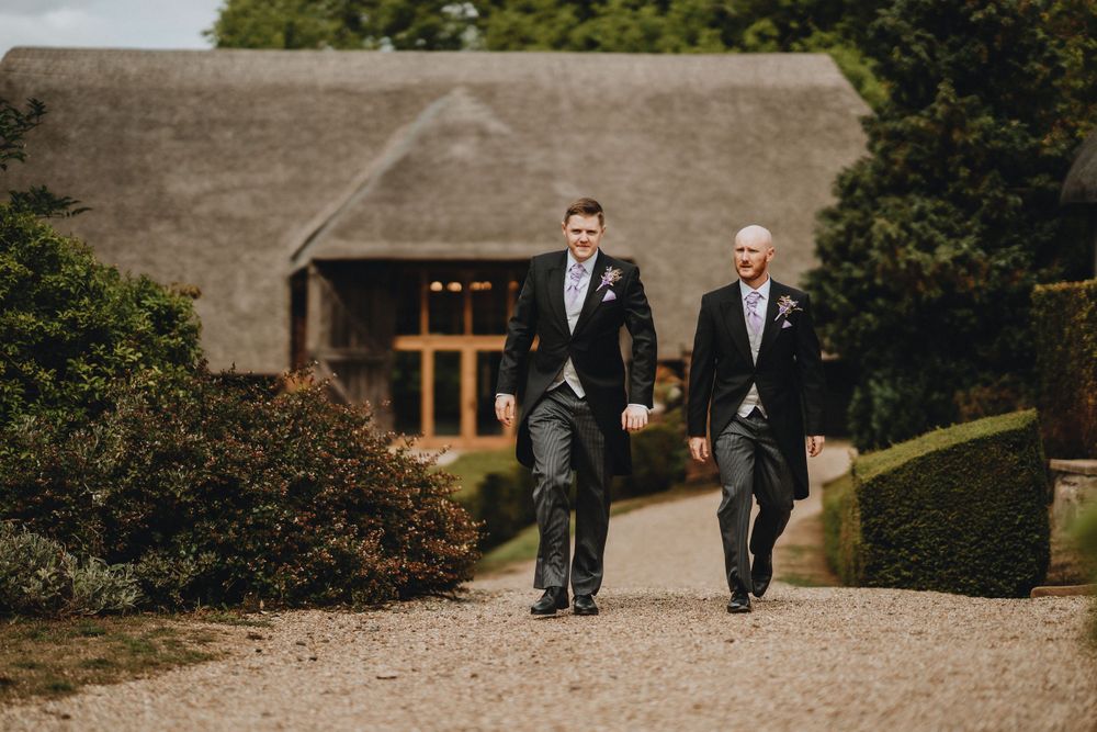 Two men in formal suits walk together on a gravel path near a rustic thatched-roof building on a cloudy day.
