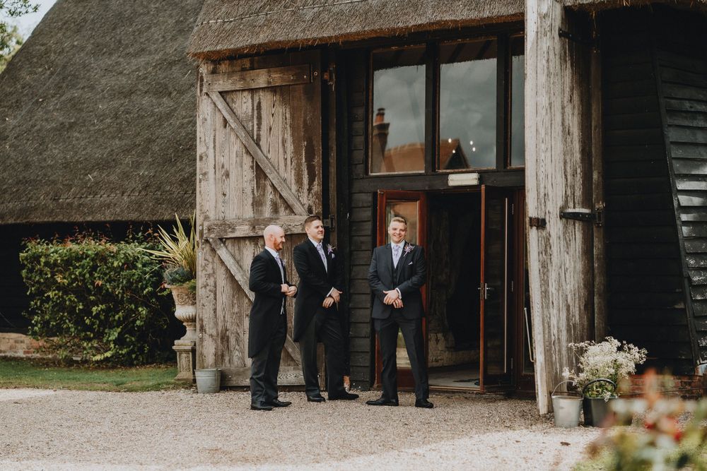 Three people in formal attire stand at the entrance of a rustic barn with thatched roof and wooden doors.