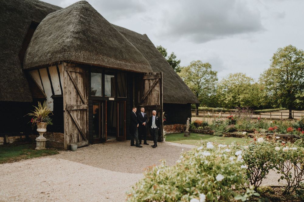 A rustic thatched-roof barn venue with wooden doors and garden landscaping on a cloudy day.