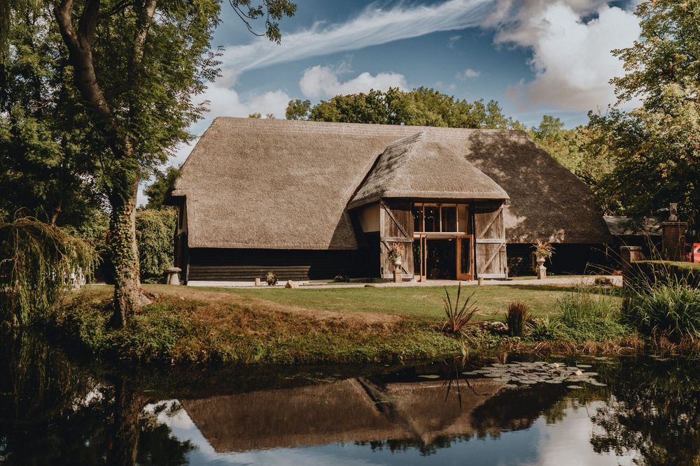 Colville Hall, rustic thatched-roof barn with wooden doors reflects in a tranquil pond surrounded by trees on a sunny day.
