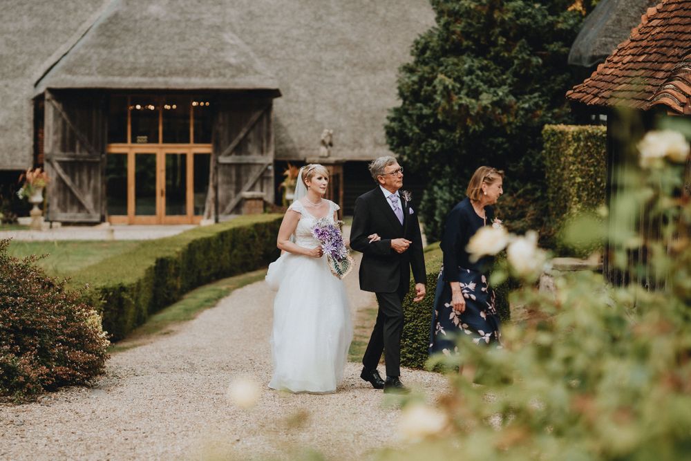 A wedding party walks along a gravel path beside a rustic barn venue with manicured hedges.