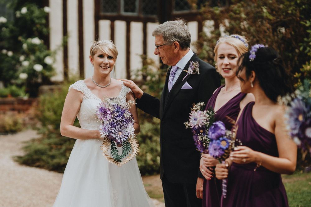 Wedding party stands outdoors holding purple and lavender bouquets during garden ceremony.