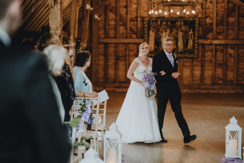 Two people walk down aisle during indoor wedding ceremony with glowing lanterns and floral decorations.