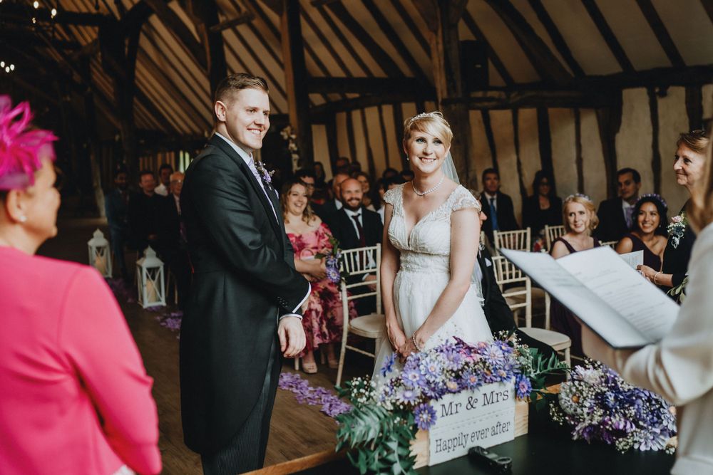 A couple stands together during their wedding ceremony in a rustic barn venue with exposed wooden beams.